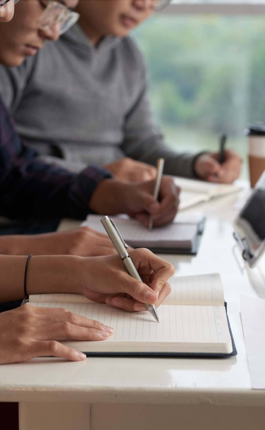 Close-up of three people writing in notebooks at a table with a window background.