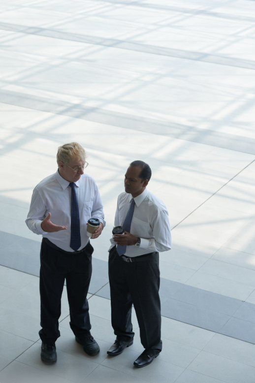 Two men in business attire having a conversation while holding coffee cups in a spacious, modern indoor setting.