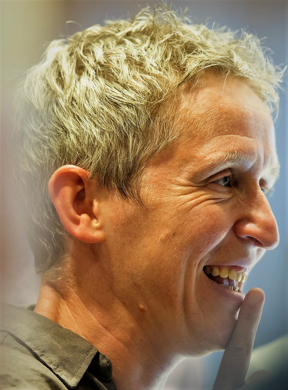 Profile of a smiling middle-aged man with short gray hair, touching his chin with his finger.